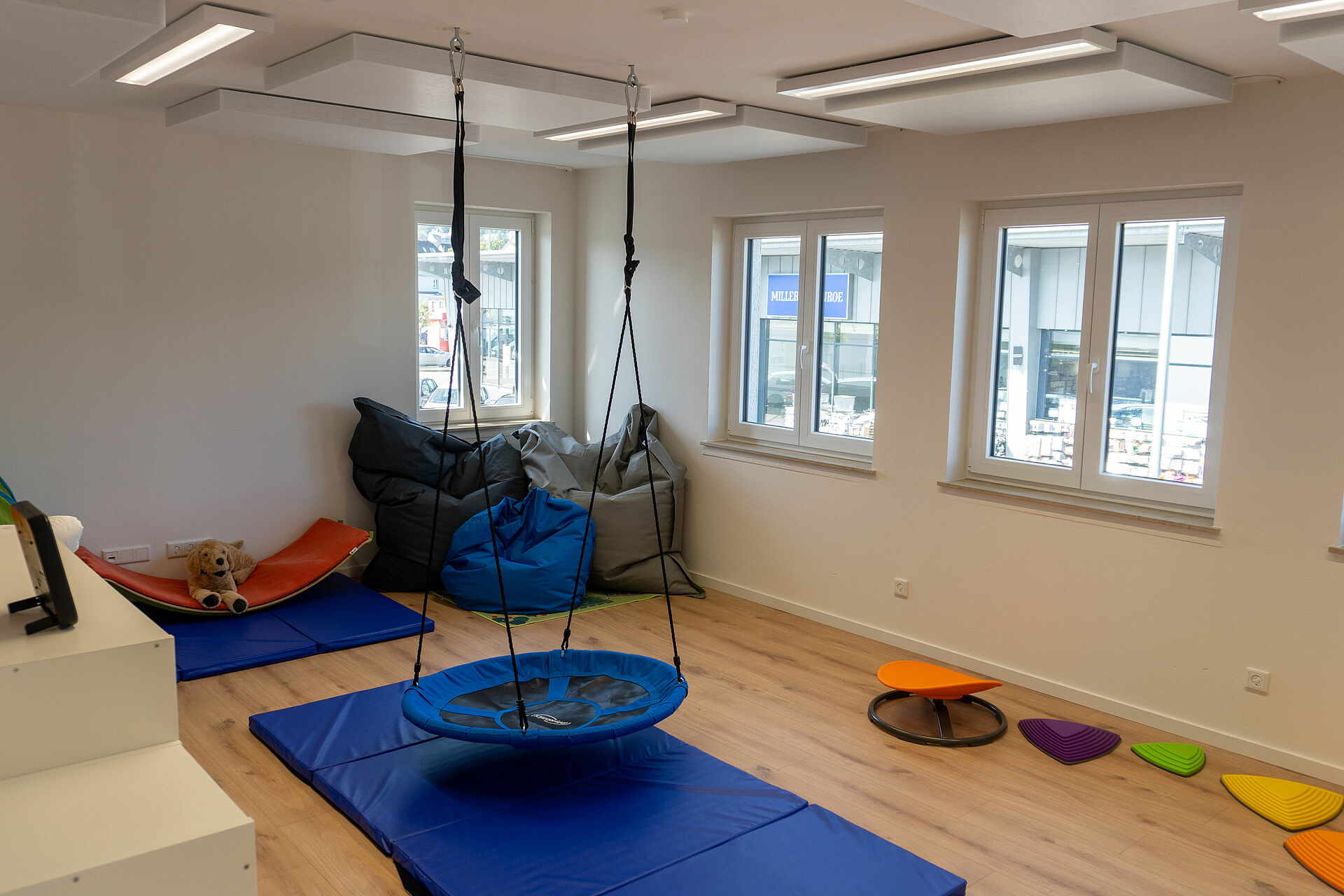 Im großen Bewegungsraum liegt zentral es eine blaue Turnmatte, über der eine Nestschaukel an der Decke hängt. Unter den Fenstern an der gegenüberliegenden Wand sind bunte Balanciersteine aufgebaut. In der Raumecke stehen drei große Sitzsäcke, daneben liegt auf einer blauen Turnmatte ein Balanceboard. Seitlich an der linken Wand ist noch ein weißes Regal zu sehen.