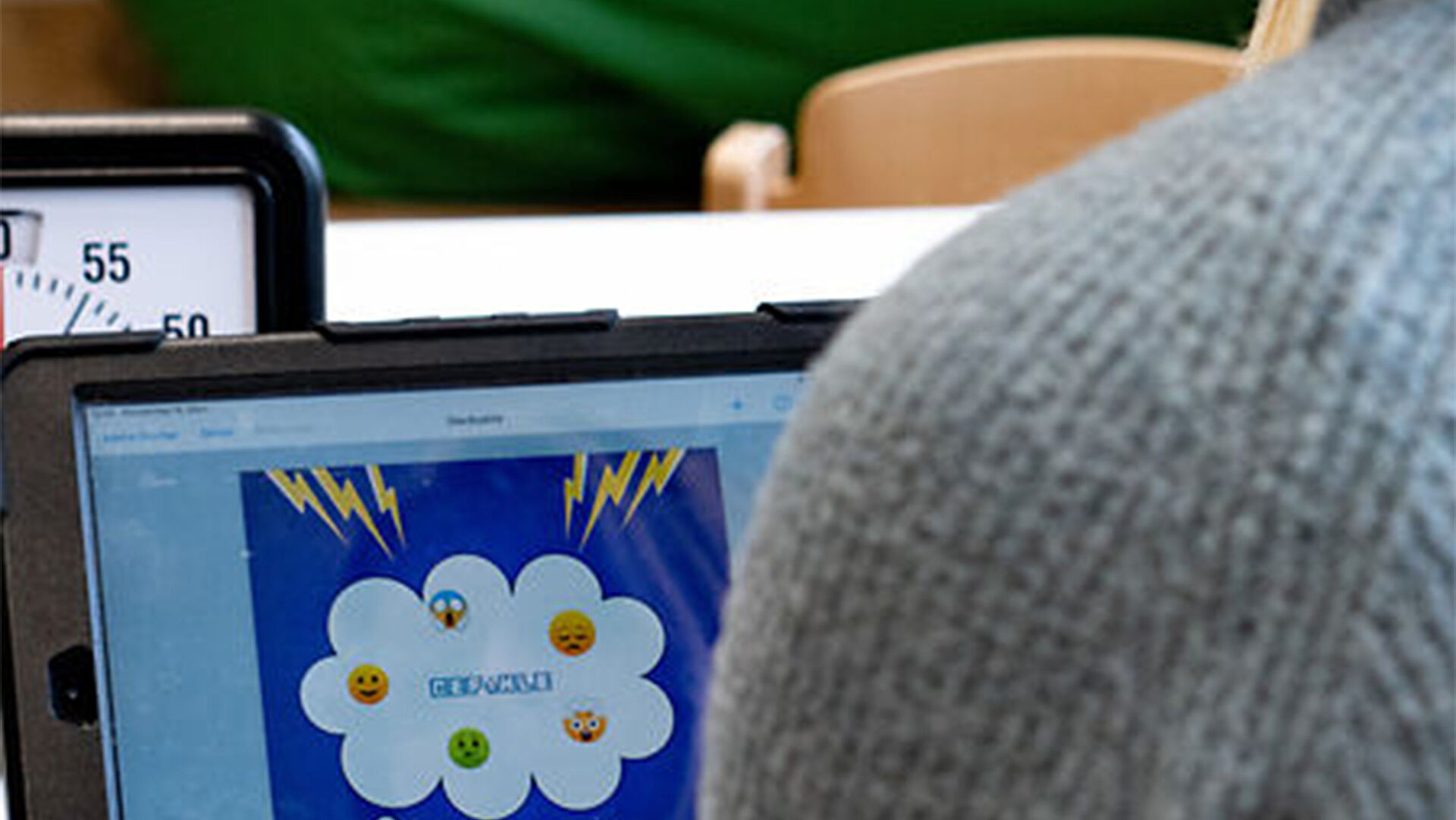 Im Vordergrund sieht man die linke Schulter eines Menschen, der vor einem Tablet sitzt. Auf dem Tablet des Bildschirms ist eine Grafik, auf der Gefühle mit Hifle von Emojis dargestellt sind.