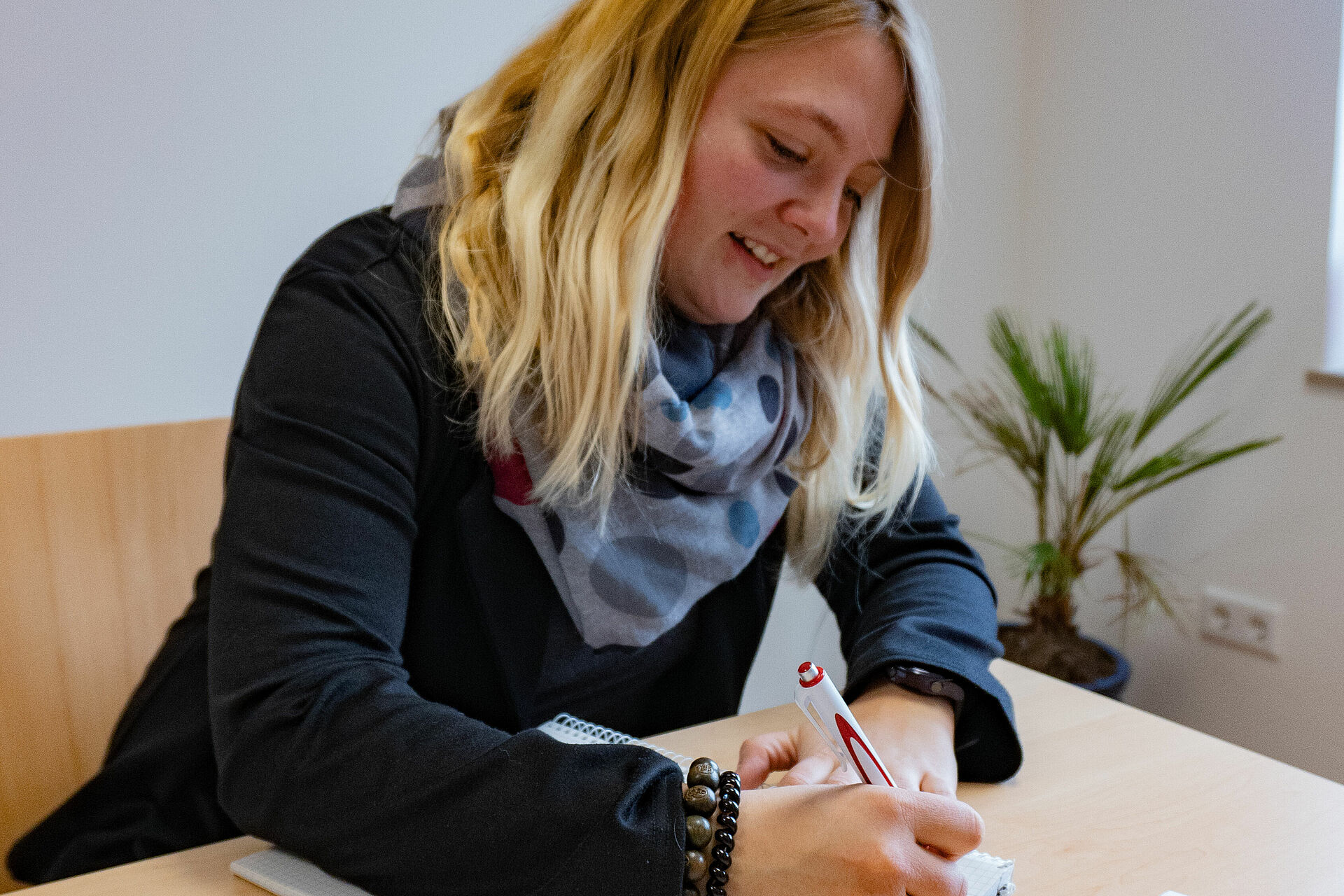 Auf dem Bild ist die Einrichtungsleitung Vera Schwindt zu sehen. Sie sitzt lachend an einem Tisch und schaut auf den vor ihr liegenden Block. In ihrer Hand hält sie einen Kugelschreiber. Sie hat blonde schulterlange Haare und trägt ein dunkelblaues Langarmshirt und einen blau gepunkteten Schal. Im Hintergrund hängt eine weiße Leinwand an der weißen Wand und auf dem Boden steht eine Pflanze.