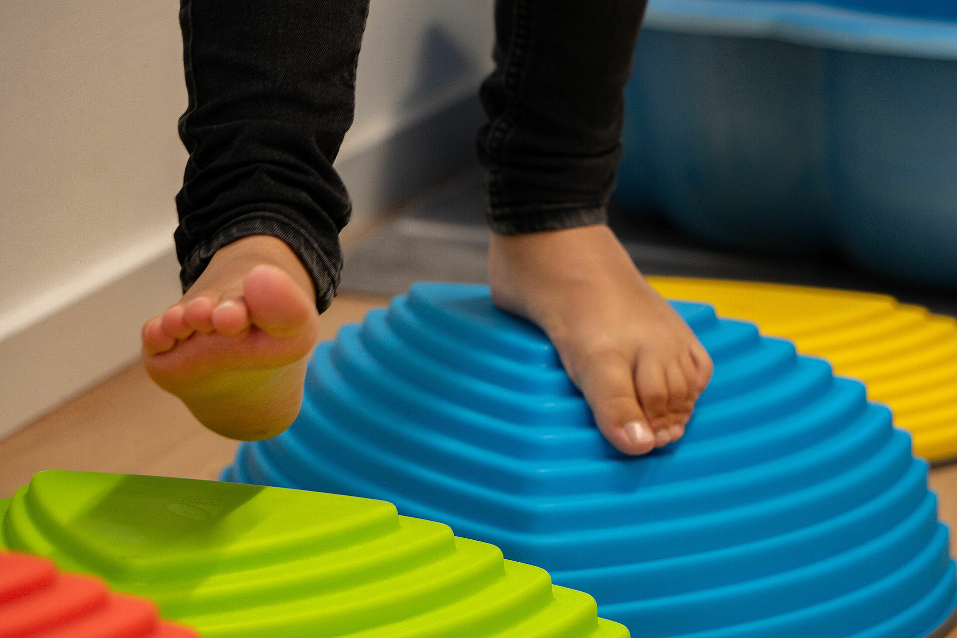 Ein Kind läuft barfuß über Balanciersteine. Die Balanciersteine sind rot, grün, blau und gelb. Es sind nur die Füße und Unterschenkel des Kindes zu sehen.