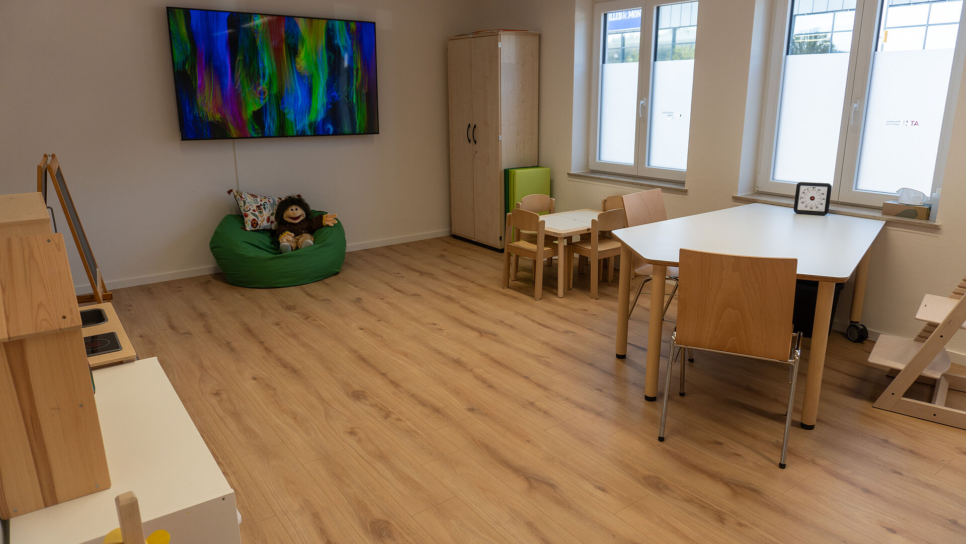 Am linken Bildrand erkennt man Möbel aus hellem Holz, gebenüber steht ein leerer Tisch mit zwei Stühlen und einem Kinderstuhl. Daneben steht noch ein kleinerer Tisch für Kinder, dahinter steht ein Schrank. An der Wand vor Kopf hängt ein abstraktes Bild in grün und blau. Darunter steht ein grünes Sitzkissen mit einem Plüschaffen. Der Boden ist aus hellem Holz und die Wände sind in hellem beige.